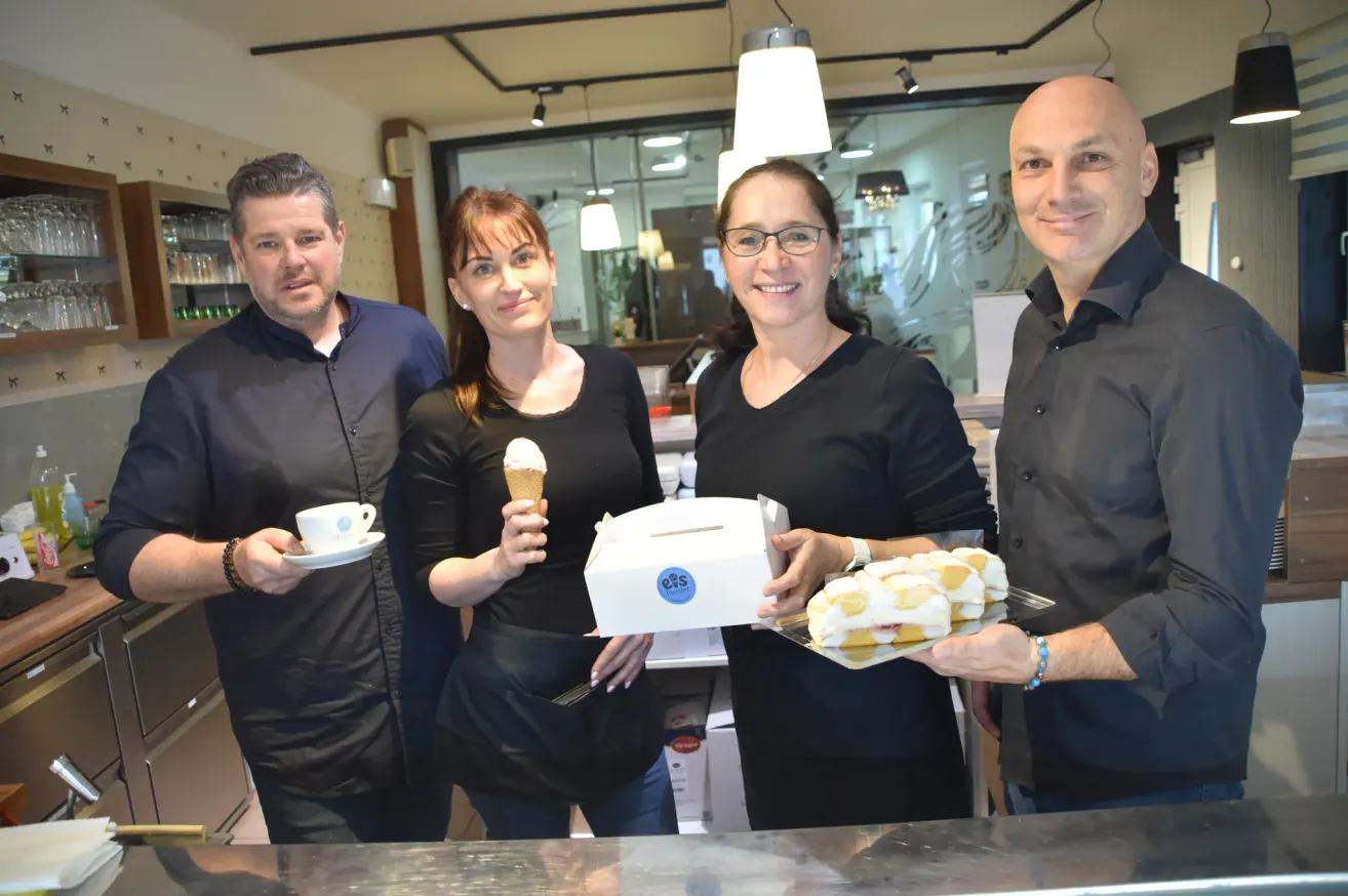 Vier Personen lächeln und posieren für ein Foto in einem Geschäft. Zwei Frauen halten ein Eis und eine Schachtel mit Sandwiches, während ein Mann rechts eine Tablett mit Sandwiches hält. Ein anderer Mann hält eine Kaffeetasse.