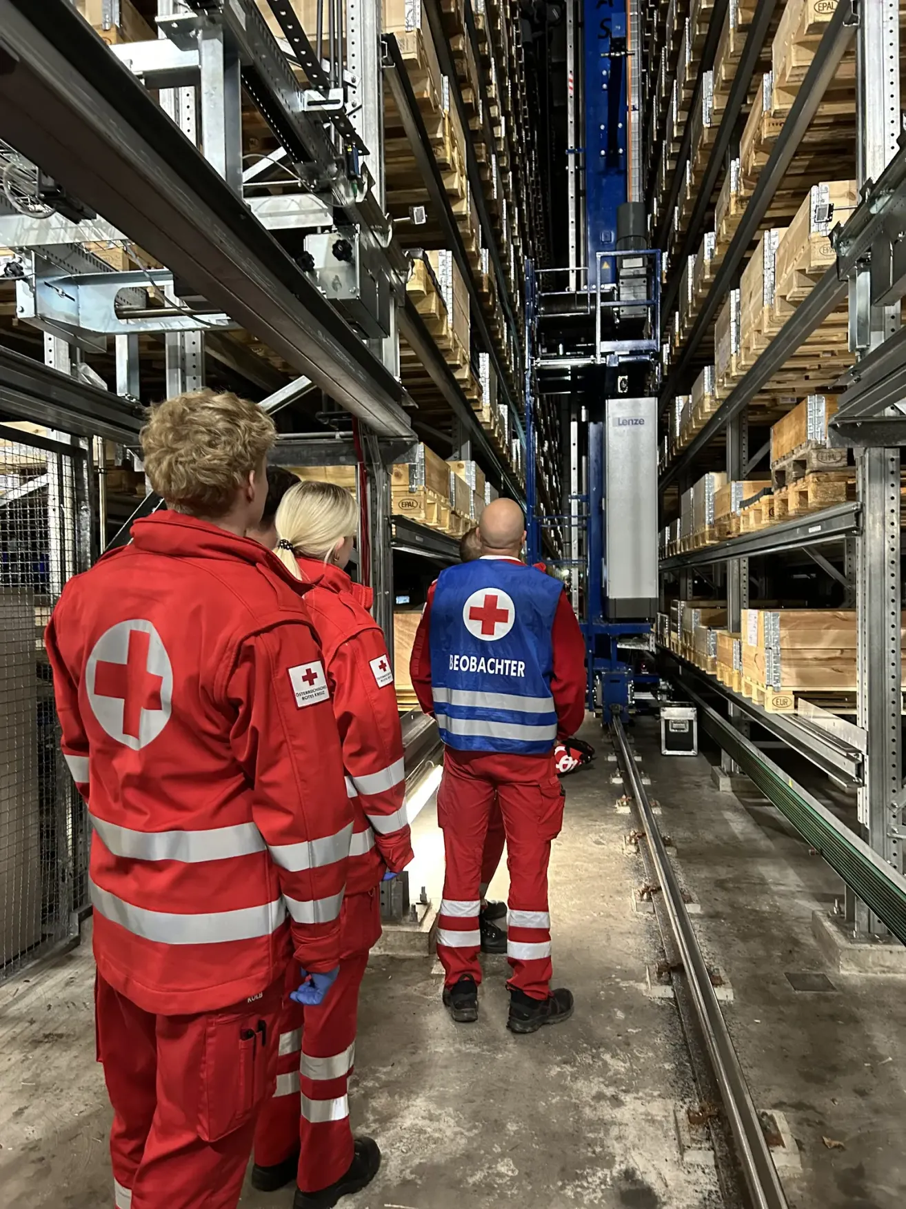 A team of medical workers in red uniforms walk through a warehouse. One wears a blue vest labeled 'Observer'. Behind them, shelves are filled with boxes.