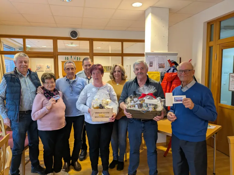 Eine Gruppe älterer Menschen versammelt sich in einem Raum, hält Geschenkboxen in der Hand und lächelt. Sie scheinen ein Ereignis zu feiern.