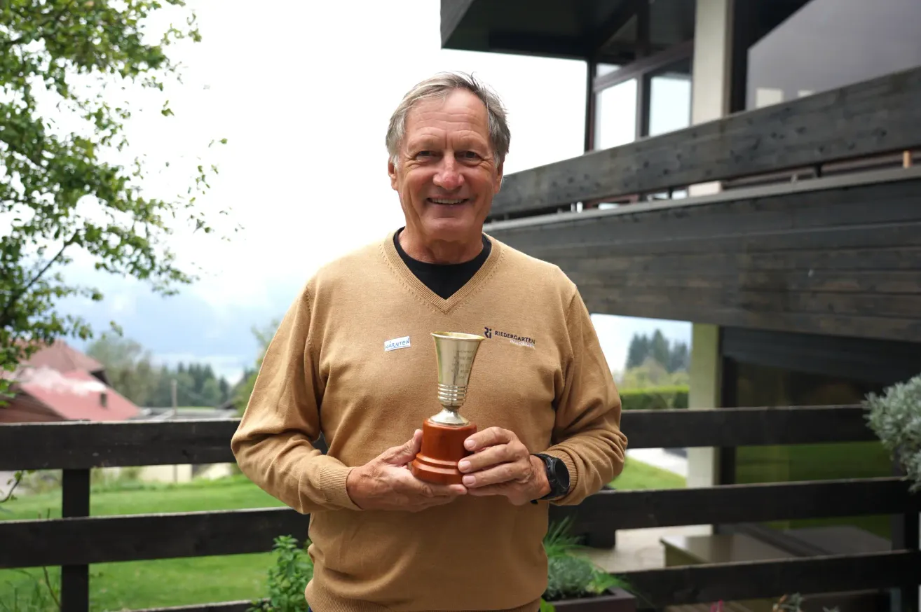 Ein älterer Mann steht draußen, lächelt und hält einen Pokal in den Händen. Er trägt einen braunen Pullover mit einem Logo auf der linken Brust. Hinter ihm befindet sich ein Holzzaun und ein Haus.