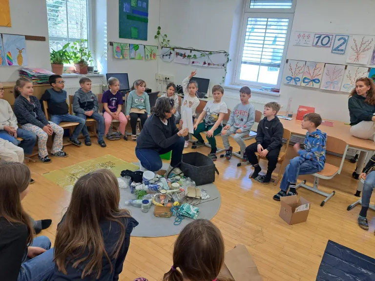 Eine Gruppe von Kindern sitzt im Kreis um eine Lehrerin, die eine Tüte mit recycelbaren Abfällen hält. Das Klassenzimmer ist gut beleuchtet und hat einen Holzboden, weiße Wände und große Fenster.