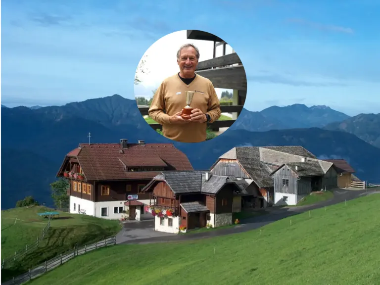 Ein Mann in einem braunen Shirt hält einen Pokal vor einem malerischen Alpendorf.