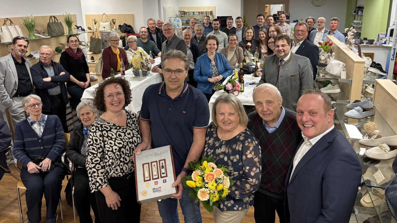 Eine Gruppe von Menschen ist in einem Raum versammelt und posiert für ein Foto. Der Mann in der Mitte hält eine Box mit einer Weinflasche darauf. Eine Frau mit einem Blumenstrauß steht neben ihm. Hinter ihnen stehen Tische mit Flaschen und anderen Gegenständen darauf. Der Raum hat weiße Wände und Holzböden.