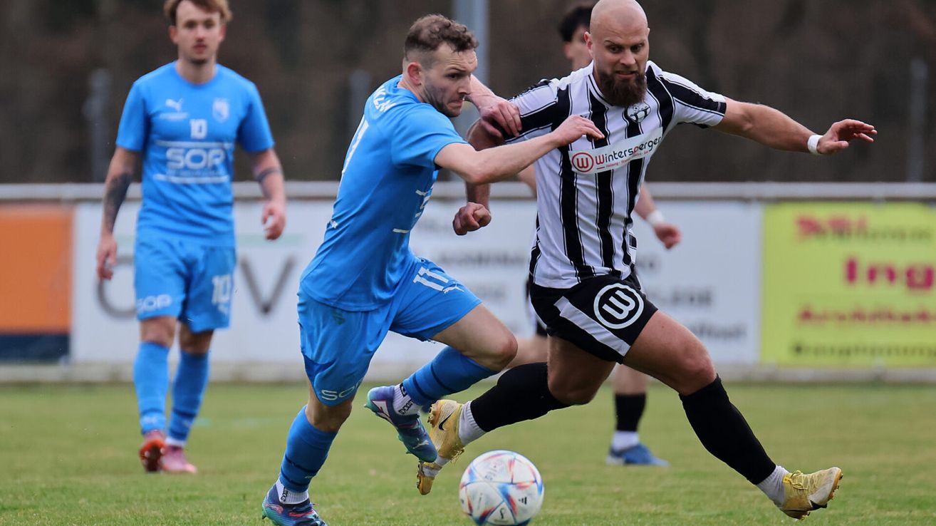 Zwei Fußballspieler sind mitten in der Aktion. Einer ist in Blau, der andere in Schwarz-Weiß gestreift. Beide laufen auf einen Fußball auf dem Feld zu.
