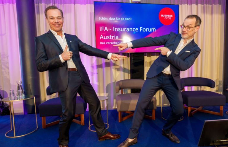 Zwei Männer in Anzügen geben im Konferenzraum den Daumen hoch. Einer zeigt auf eine große Leinwand mit dem Text 'IFA - Versicherungsforum Österreich'.