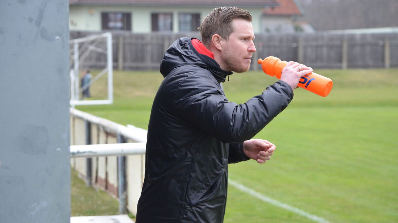 Ein Mann in einer schwarzen Jacke steht auf einem Fußballfeld und hält eine orangefarbene Wasserflasche. Er sieht konzentriert aus und ist möglicherweise Trainer oder Ausbilder.