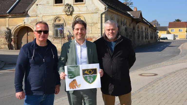 Drei Männer stehen auf einer Straße, einer hält ein Schild. Der Mann links trägt eine Sonnenbrille und eine blaue Jacke. Der mittlere Mann trägt einen grünen Blazer und ein weißes Hemd und hält ein Schild mit einem Mammut-Illustration. Der Mann rechts trägt eine schwarze Jacke. Hinter ihnen ein altes Gebäude mit Bogenfenstern und einer Steinfassade.