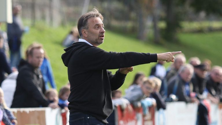 Ein Trainer leitet ein Fußballspiel mit erhobenem Arm, trägt eine schwarze Kapuzenjacke und Jeans, während Zuschauer im Hintergrund zuschauen.
