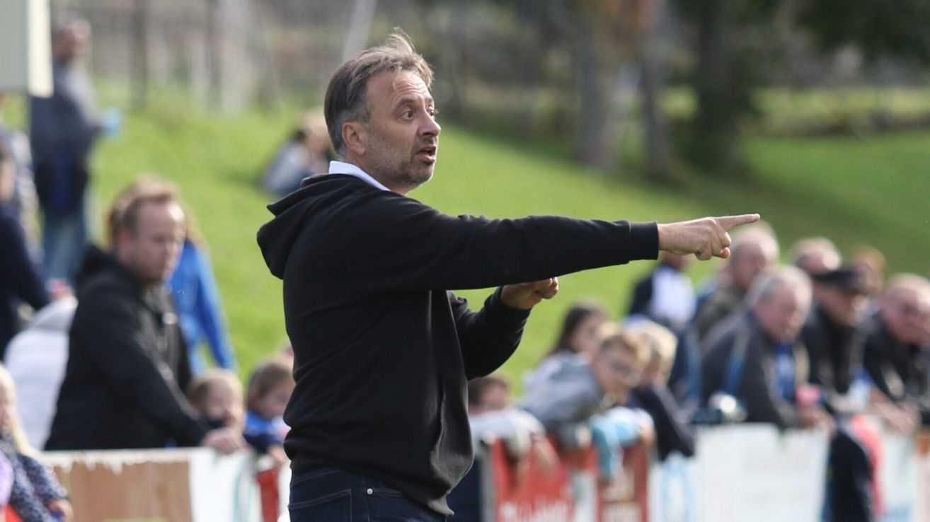 Ein Trainer leitet ein Fußballspiel mit erhobenem Arm, trägt eine schwarze Kapuzenjacke und Jeans, während Zuschauer im Hintergrund zuschauen.