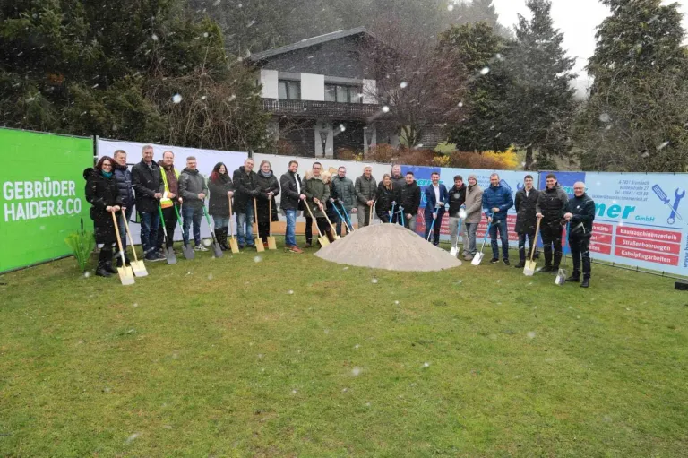 A group of people holding shovels stand around a pile of dirt in a snowy field. Behind them is a banner and a building.