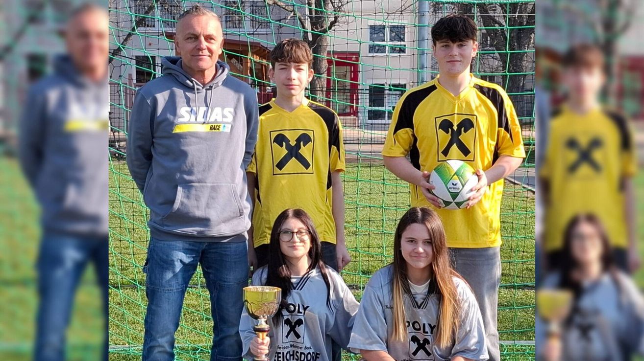 A group of four young people pose for a photo with a soccer ball and trophy. They wear matching jerseys with a logo. The man in the gray hoodie stands behind them. The background features a soccer net and a building.