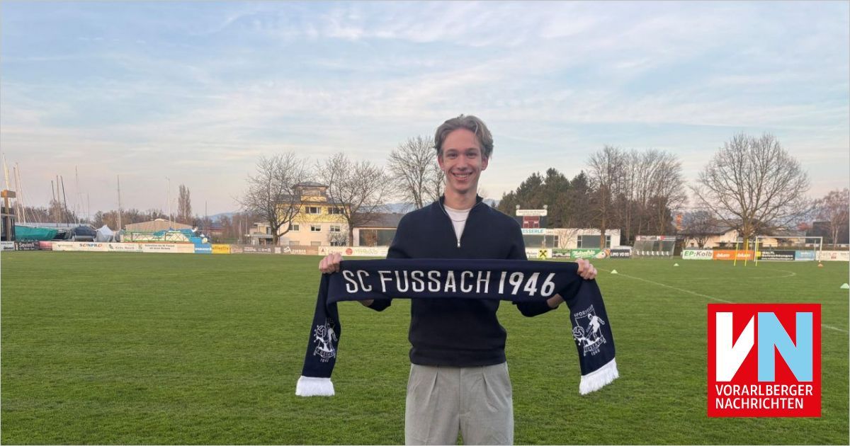 Ein Mann steht auf einem Rasenfeld und hält ein Banner mit der Aufschrift 'SC FUSSACH 1946' in der Hand, lächelt mit einem Stadion im Hintergrund.