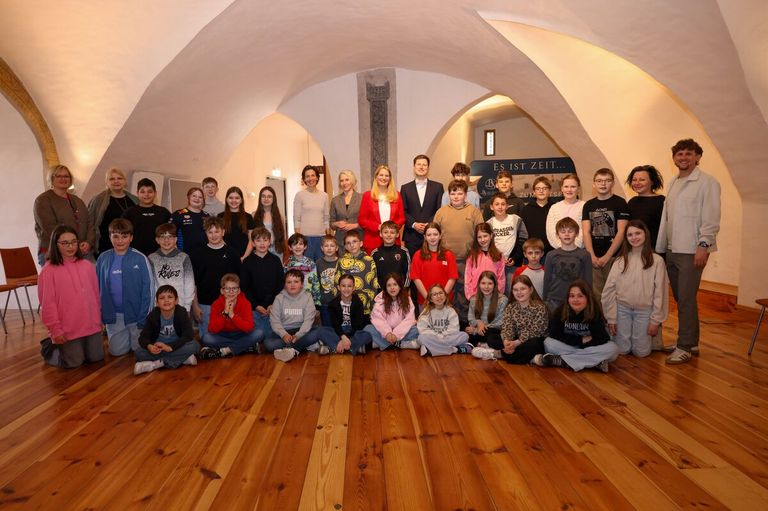Eine Gruppe von Schülern und Lehrern posiert für ein Foto in einem Raum mit Holzböden und gewölbten Decken. Einige tragen Brillen und Jacken. Ein Schild steht für 'Es ist Zeit...'.