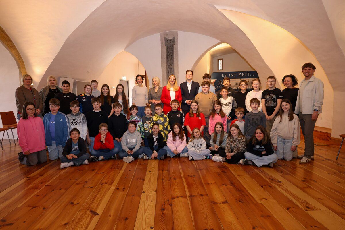 Eine Gruppe von Schülern und Lehrern posiert für ein Foto in einem Raum mit Holzböden und gewölbten Decken. Einige tragen Brillen und Jacken. Ein Schild steht für 'Es ist Zeit...'.