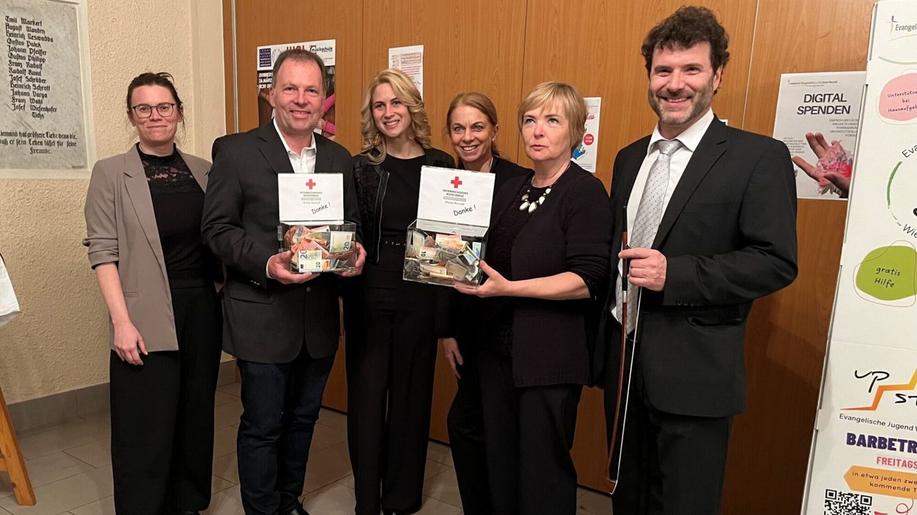 Fünf Personen halten Boxen mit Geld. Sie lächeln und schauen in die Kamera. Auf den Boxen ist ein rotes Kreuz. Hinter ihnen hat eine Wand Poster darauf.