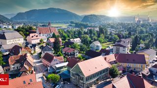 Ein Luftbild eines malerischen Dorfes mit bunten Häusern und einer Kirche, umgeben von Bäumen und Bergen unter einem untergehenden Sonnenlicht.