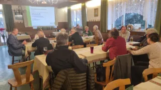 In einem Raum mit weißen Wänden sitzen mehrere Personen um einen Tisch herum. Sie konzentrieren sich auf eine Präsentation an der Projektionsleinwand. Der Tisch ist mit einer weißen Tischdecke bedeckt.