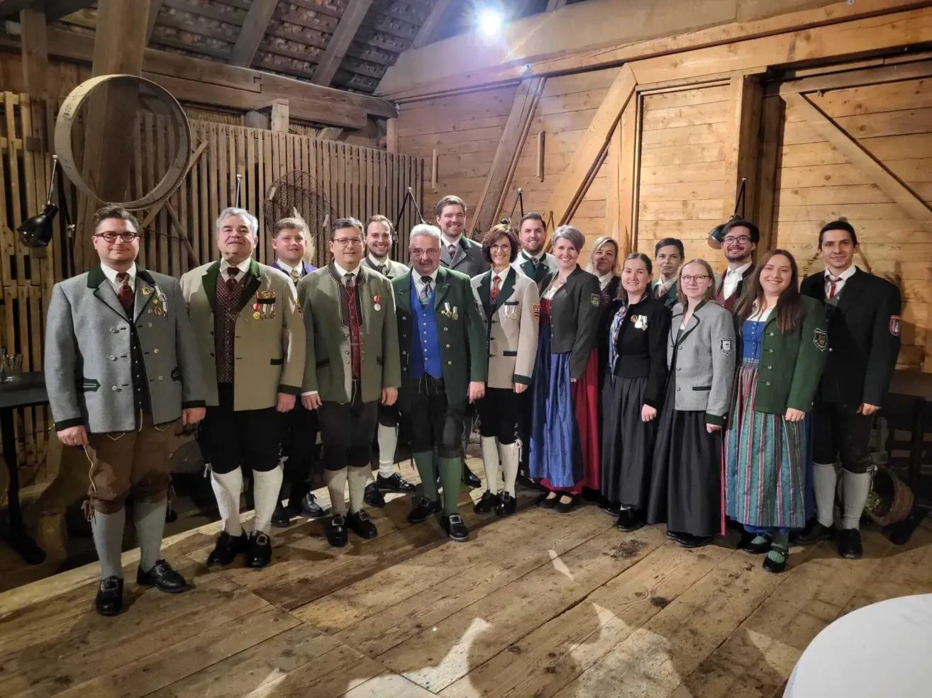 Eine Gruppe von Menschen in traditioneller Kleidung posiert für ein Foto in einem Holzschuppen. Sie stehen in einer Reihe mit Holzwänden und einer Metallstruktur im Hintergrund.