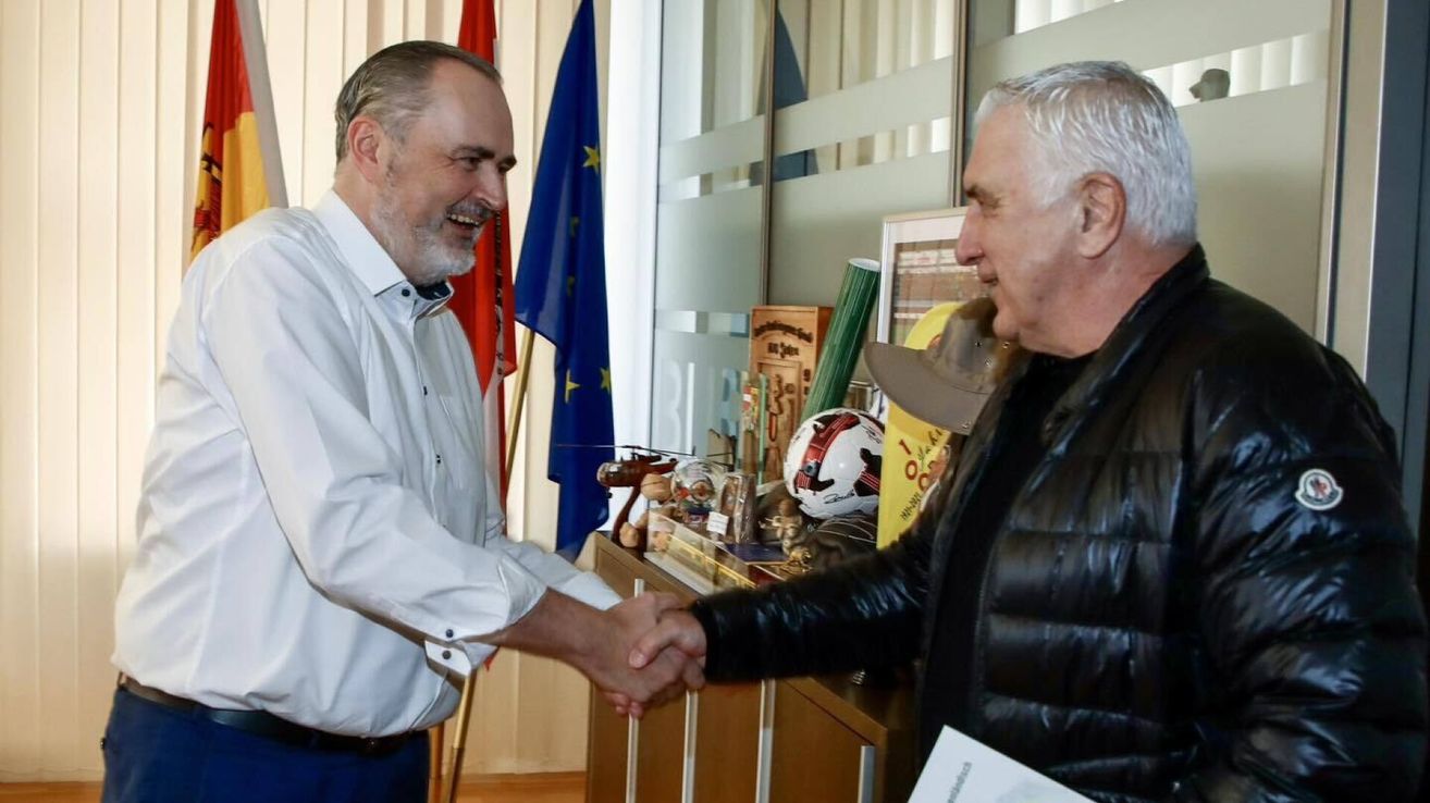 Zwei Männer schütteln sich die Hände in einem Büro. Hinter ihnen stehen Flaggen, Trophäen und Bücher auf einem Schreibtisch. Ein Mann trägt ein weißes Hemd, der andere einen schwarzen Mantel.