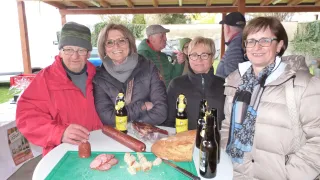 Eine Gruppe von Erwachsenen steht um einen Tisch mit Brot, Bierflaschen und Fleisch. Sie lächeln und scheinen für ein Foto zu posieren.