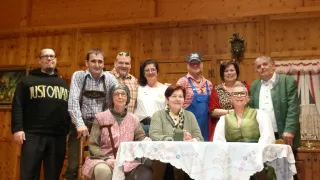 Eine Gruppe von Menschen posiert für ein Foto in einer Inneneinrichtung. Sie sind in verschiedenen Kleidungsstücken gekleidet und einige von ihnen sitzen auf Stühlen. Vor ihnen steht ein Tisch. Hinter ihnen ist eine Holzwand und eine Lampe hängt an der Wand.