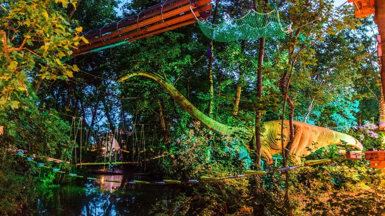 Eine gelbe Dinosaurierstatue steht in einem Wald bei Dämmerung, beleuchtet von bunten Lichtern, mit einer Seilbrücke darüber.