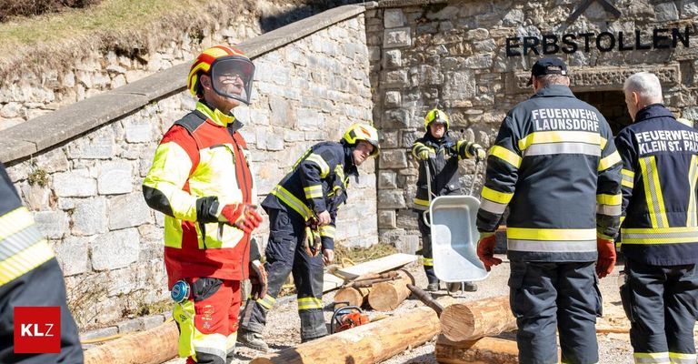 Feuerwehrleute in Schutzkleidung arbeiten mit Werkzeugen und Baumstämmen an einem Projekt. Eine Wand mit dem Wort 'ERBS' ist hinter ihnen zu sehen.