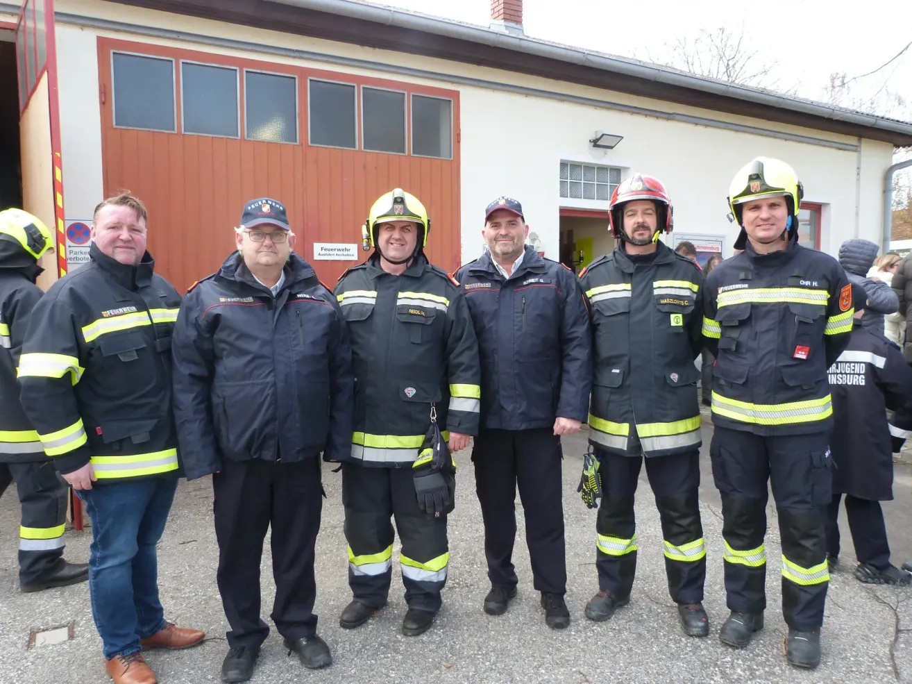 Eine Gruppe von Feuerwehrleuten in Uniform steht vor einem Feuerwehrhaus. Die Feuerwehrleute lächeln und das Gebäude hat eine rote Tür und Glasfenster.