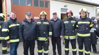 Eine Gruppe von Feuerwehrleuten in Uniform steht vor einem Feuerwehrhaus. Die Feuerwehrleute lächeln und das Gebäude hat eine rote Tür und Glasfenster.