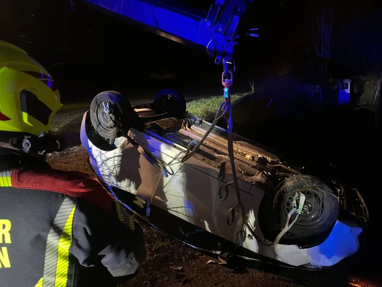 Ein umgekipptes Fahrzeug wird von einem Kran mit einem Gurtzeug angehoben, das an einem Vorderrad befestigt ist. Das Auto befindet sich auf einer Grasfläche, und eine Person ist in der Nähe.