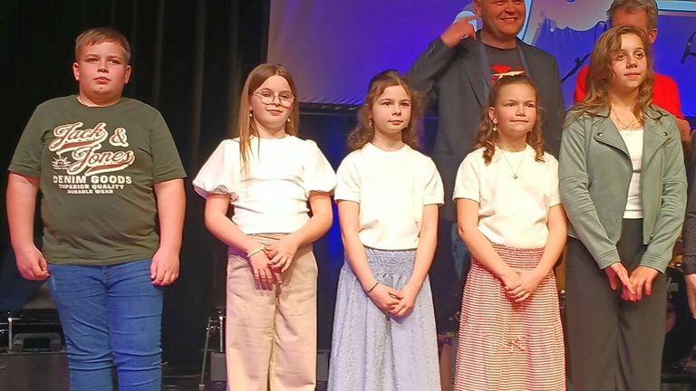 Drei Mädchen in weißen Oberteilen stehen auf der Bühne mit einem Mann im Anzug hinter ihnen. Sie alle schauen nach vorne.