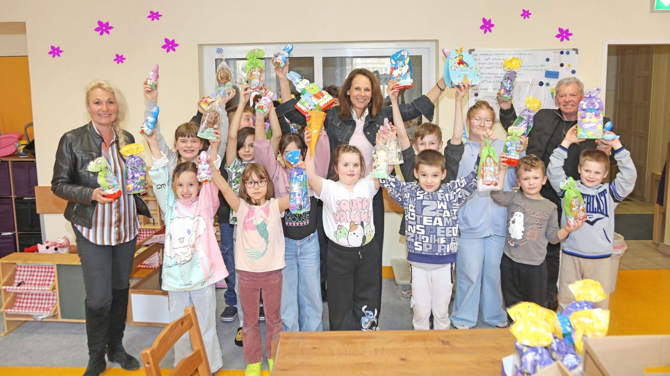 Children and a woman holding Easter eggs in a classroom, smiling, and posing for a photo. The woman is standing in front, and the children are raising their hands with various Easter eggs.