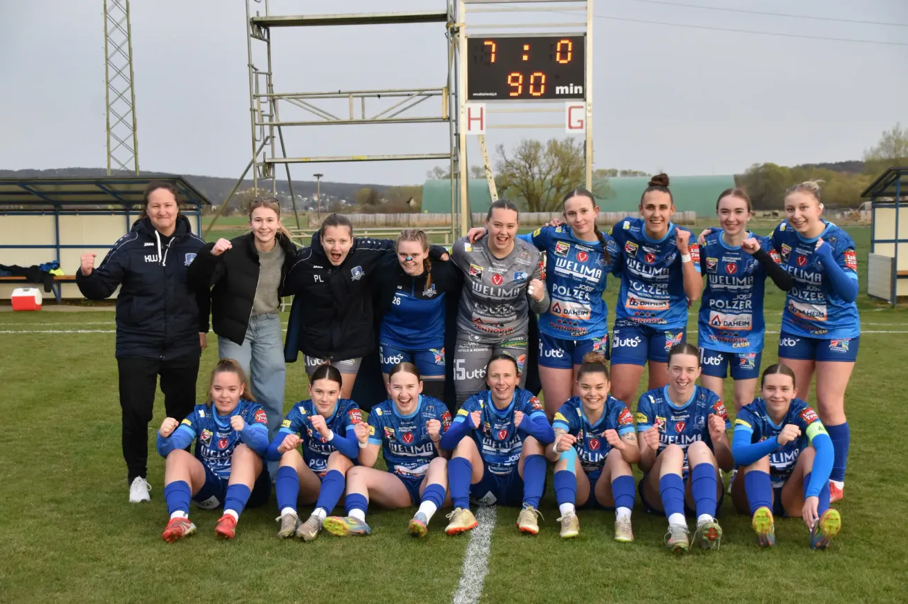 Eine Frauensportmannschaft posiert für ein Foto mit einem Anzeigetafel im Hintergrund, die einen Score von 7:0 anzeigt. Die Mannschaft trägt einheitliche Uniformen.