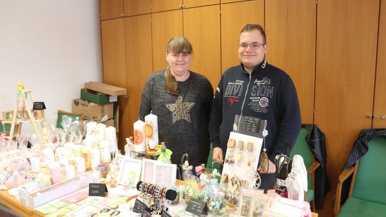 Zwei Personen stehen hinter einem Tisch mit verschiedenen Handwerksarbeiten und lächeln. Einer trägt eine Brille und eine schwarze Jacke mit Text, der andere ein graues Shirt mit einem Stern. Dahinter stehen Holzschränke und Kisten. Der Tisch hat Kerzen, Armbänder und Karten.