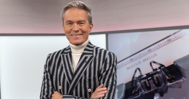 Ein Mann in einer gestreiften Jacke und weißem Rollkragen steht lächelnd und mit verschränkten Armen vor einem Bildschirm, auf dem eine Handtasche beworben wird.