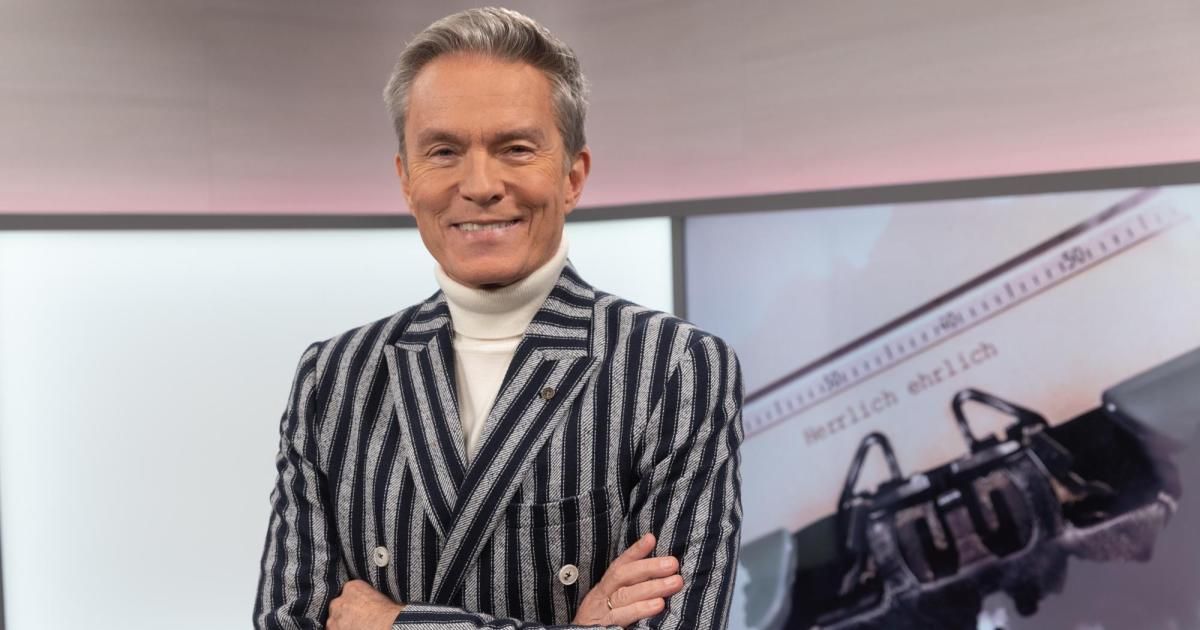 Ein Mann in einer gestreiften Jacke und weißem Rollkragen steht lächelnd und mit verschränkten Armen vor einem Bildschirm, auf dem eine Handtasche beworben wird.
