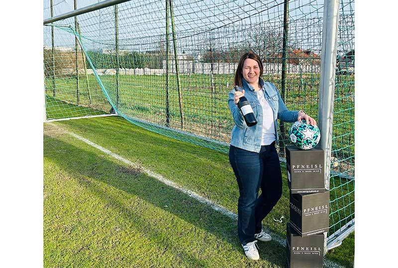 Eine Frau steht auf einem Fußballfeld und hält eine Flasche und einen Fußball. Hinter ihr steht ein Fußballtor mit einem Netz.