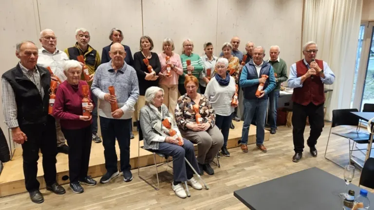 A group of people, possibly seniors, are standing and sitting in a room, holding orange packages. They are smiling and seem to be posing for a photo. They are all wearing glasses and sneakers. Some of them are holding a stick. The room has a wooden floor and white walls.