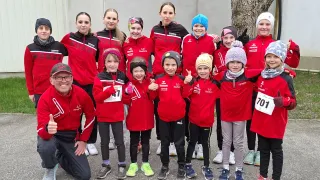 Eine Gruppe junger Läufer und Trainer in roten Jacken posiert für ein Foto. Einige haben Nummern auf ihren Trikots, und sie tragen Mützen und Turnschuhe. Der Hintergrund besteht aus einem Gebäude und einem Baum.