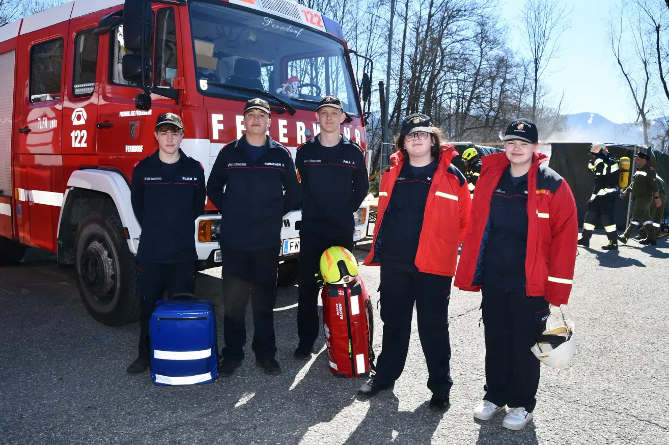 Fünf junge Feuerwehrleute stehen vor einem Feuerwehrwagen. Sie tragen Uniformen und Helme. Ein roter Rucksack und ein blauer Koffer liegen auf dem Boden.