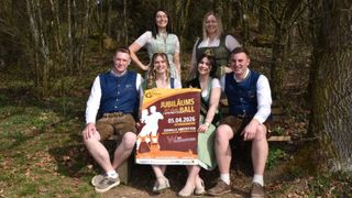 Eine Gruppe von Menschen, gekleidet in traditioneller Kleidung, posiert für ein Foto im Wald. Eine Frau in der Mitte hält ein Banner, das einen Jubilaums Ball am 5. April 2026 ankündigt.