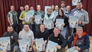 Eine Gruppe von Menschen, die Urkunden halten, lächeln und für ein Foto posieren. Sie stehen vor einem roten Vorhang. Einige tragen Jacken und Brillen. Sie halten Urkunden mit Namen und Veranstaltungsdetails.