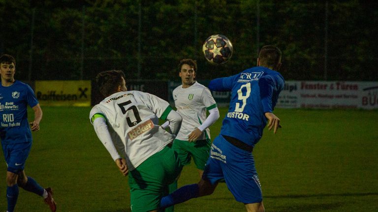 Drei Fußballspieler konkurrieren auf dem Spielfeld. Der Spieler in Blau versucht, den Ball zu köpfen. Der Spieler in Grün versucht zu blocken, während ein anderer Spieler in Weiß zusieht.