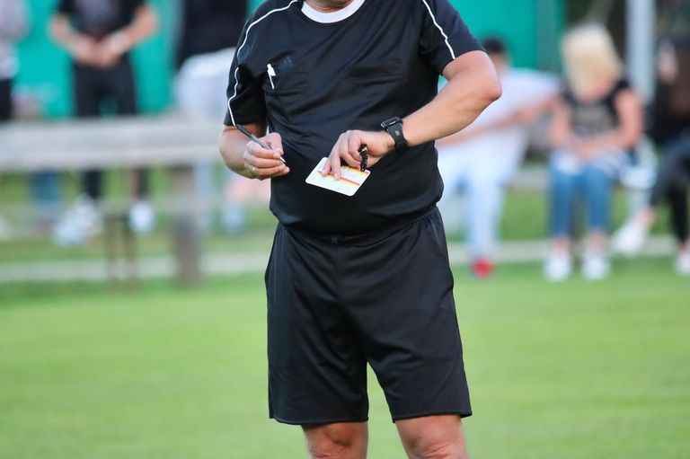 Ein Fußballschiedsrichter hält eine Karte und einen Stift auf einem Rasenfeld und trägt ein schwarzes Trikot mit weißen Streifen.