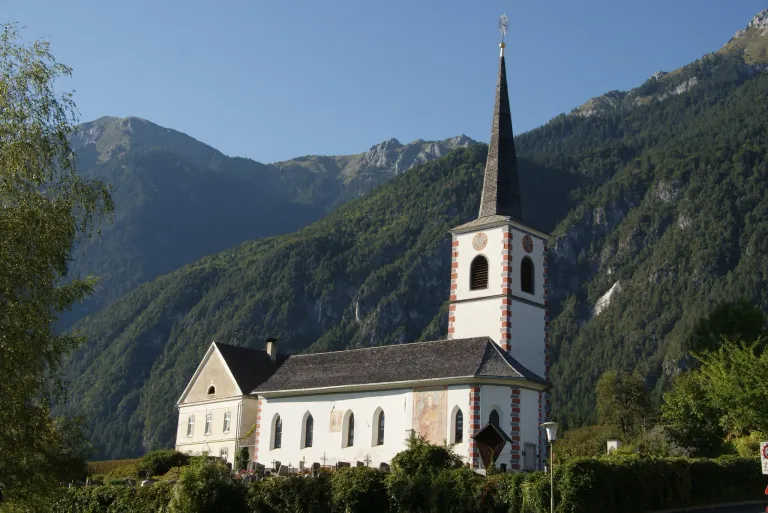 Eine weiße Kirche mit einem Turm steht vor einer Bergkulisse. Die Kirche hat ein graues Dach, viele Fenster und eine Wandmalerei an der Seite. Der Himmel ist klar und blau.