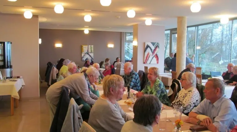 Mehrere ältere Menschen sitzen an einem Tisch in einem Restaurant. Einige unterhalten sich, andere schauen sich um. Eine Frau trägt eine Brille.