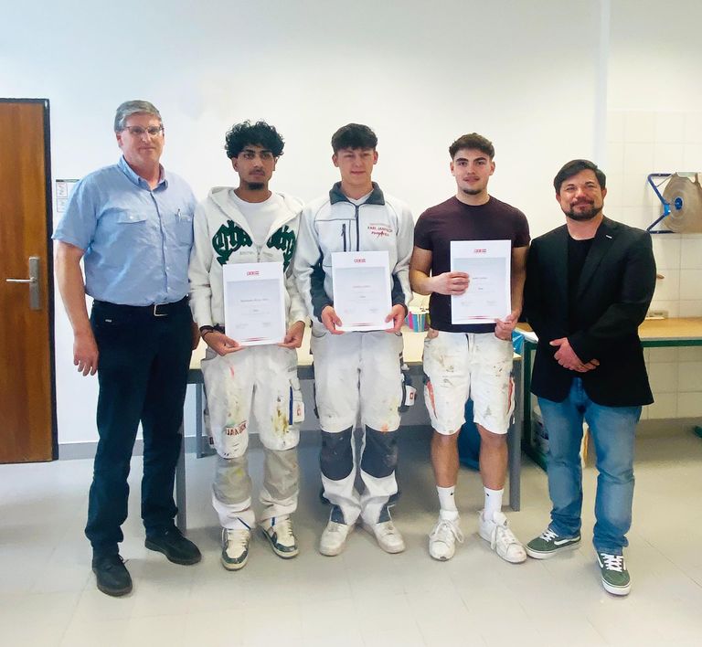 Fünf Männer stehen in einem Raum und halten Urkunden in den Händen. Die Männer tragen farbenbeschmutzte Kleidung. Dahinter ist ein Tisch mit einigen Gegenständen zu sehen.