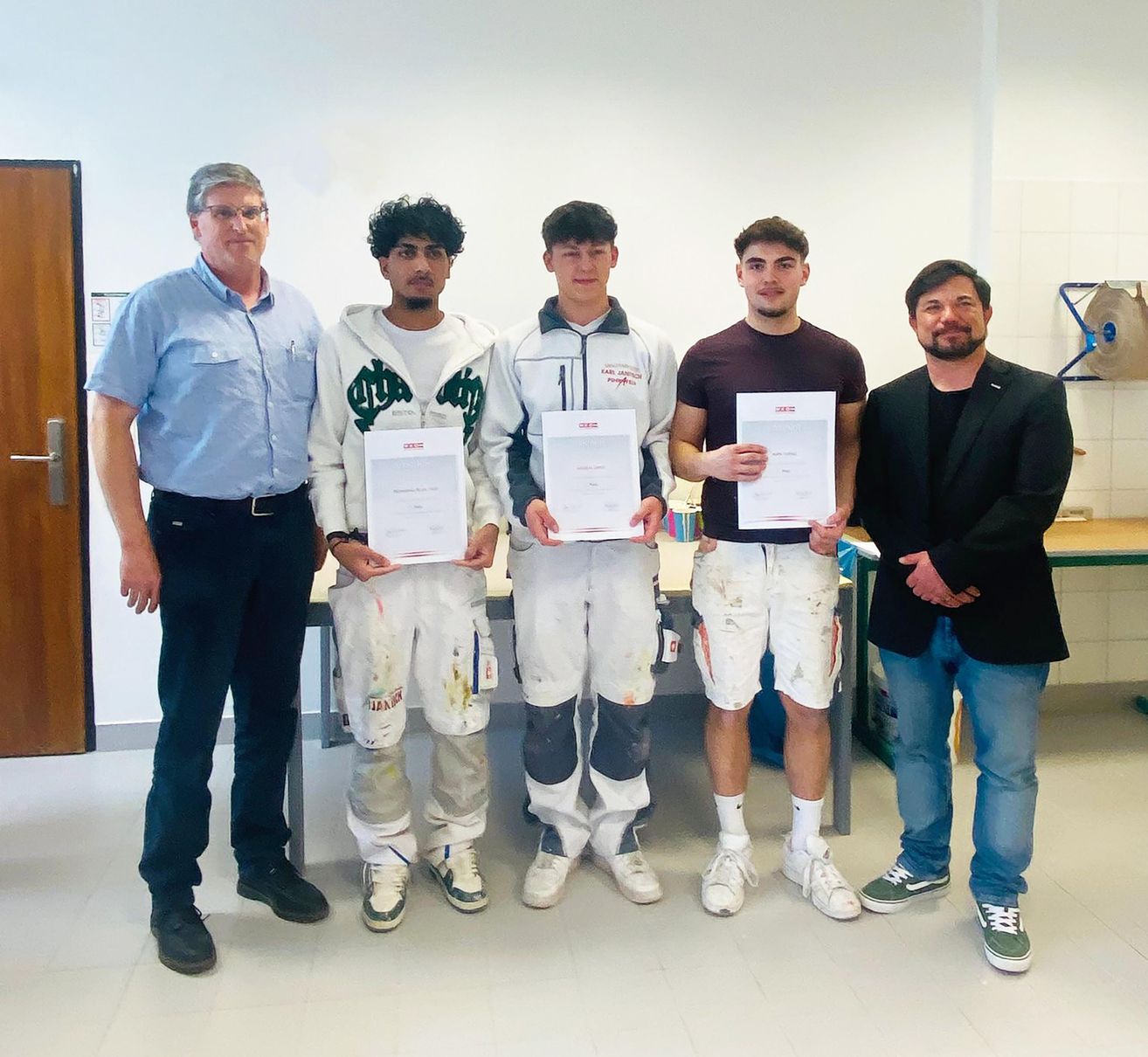 Fünf Männer stehen in einem Raum und halten Urkunden in den Händen. Die Männer tragen farbenbeschmutzte Kleidung. Dahinter ist ein Tisch mit einigen Gegenständen zu sehen.