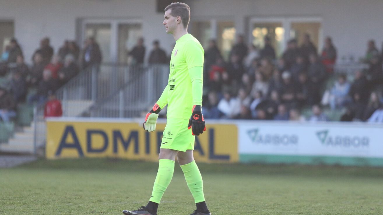 Ein Fußballspieler in neongrüner Ausrüstung steht auf einem Feld, mit dem Blick nach links, während die Zuschauer im Hintergrund verschwommen sind.
