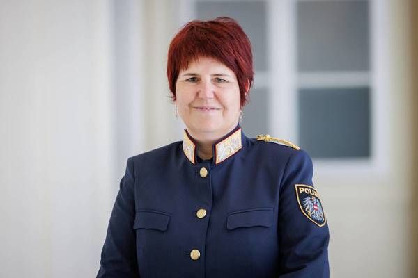Eine Frau mit roten Haaren, die eine dunkelblaue Uniform mit goldenen Knöpfen trägt, steht vor einer weißen Wand mit einem verschwommenen Fenster dahinter.